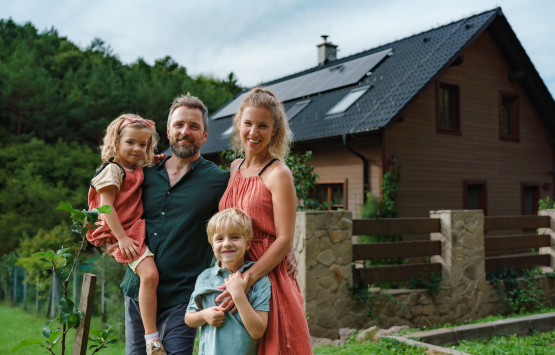 Family of four standing outside their home with rooftop solar panels surrounded by green trees.