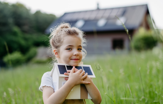 Young girl holding a model solar-powered house in a grassy yard with a solar-equipped home in the background.